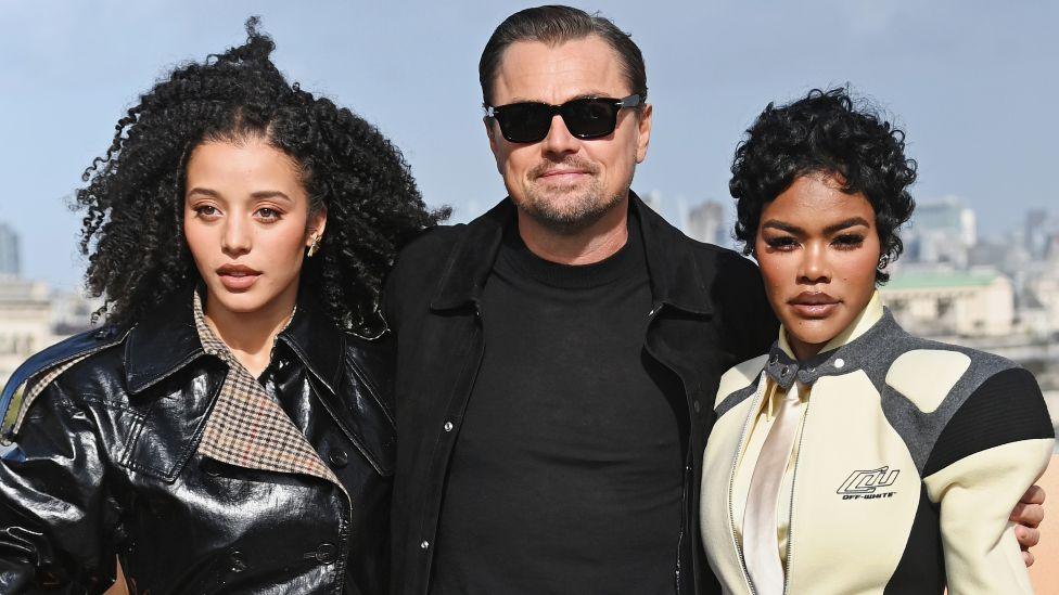 Chase Infiniti, Leonardo DiCaprio dhe Teyana Taylor marrin pjesë në fotothirrjen në Londër për One Battle After Another në Londër në shtator. Infiniti ka veshur një xhaketë të zezë me shkëlqim, DiCaprio ka veshur një xhaketë dhe bluzë të zezë dhe Taylor ka veshur një xhaketë krem, gri dhe të zezë në stilin e garave.
