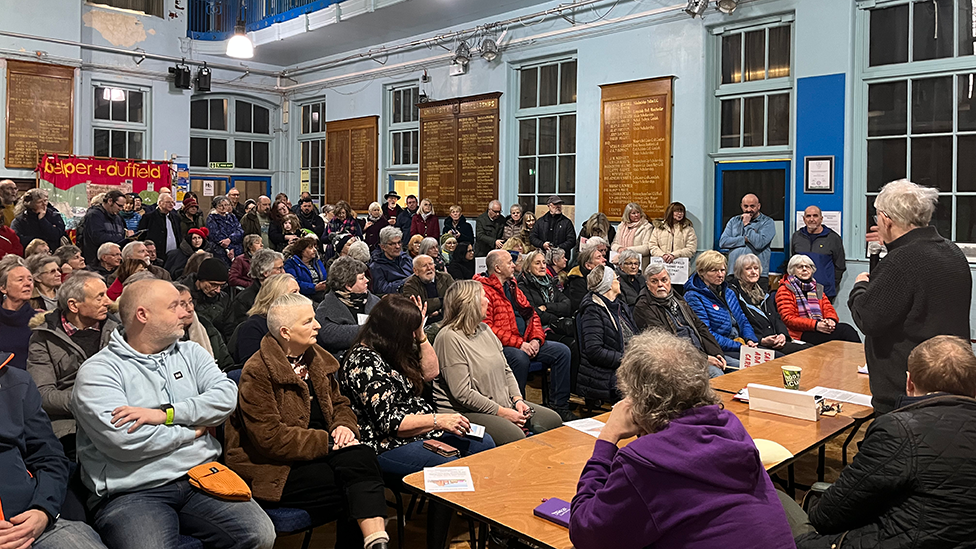 A packed Strutts Community Centre, where campaigners met with councillors and the local MP. People sit in rows of chairs as three people behind a desk answer questions