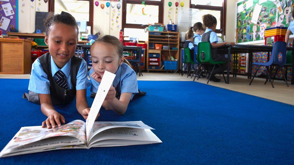 Two children lie on blue carpet in a classroom floor, reading a book together 