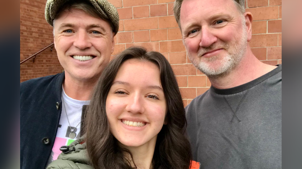Selfie of Nancy, Jonny and her dad. All three are smiling at the camera. Nancy is in the middle and has dark brown long hair. Jonny is on the left and is wearing a cap, white t-shirt and black jacket. Her dad, who has light hair and beard, is wearing a grey jumper.