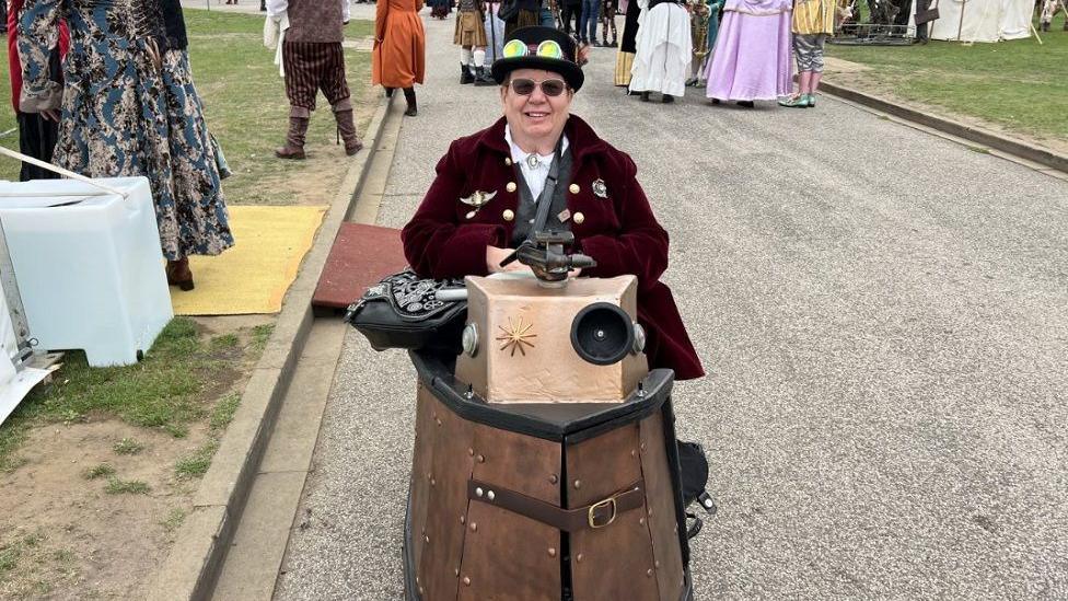 A woman sits in what appears to be a wooden boat, in a barrel shape, wearing a burgundy jacket with badges on and a black hat with sunglasses. 