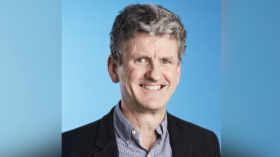 Photo of Professor Miles Parkes against a blue background. He is wearing a back jacket and blue check shirt. He is smiling at the camera.