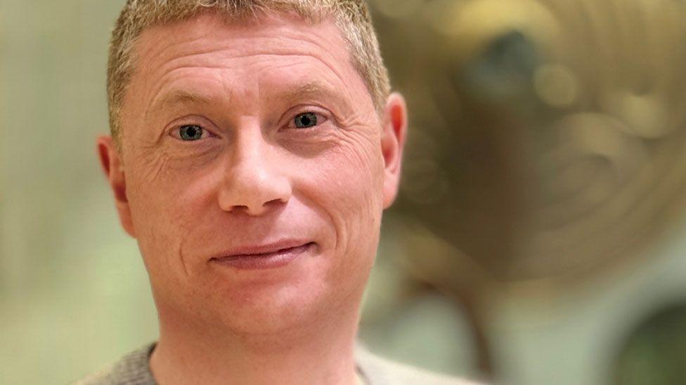 A close-up of Dr Jody Joy. He has close cropped fair hair and is clean shaven. He is wearing a grey jumper and is looking towards the camera as if about to smile.