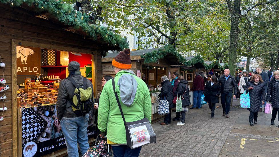 York Christmas market with a series of wooden huts lined up on the left all displaying Christmas-themed items for sale with a number of people walking past