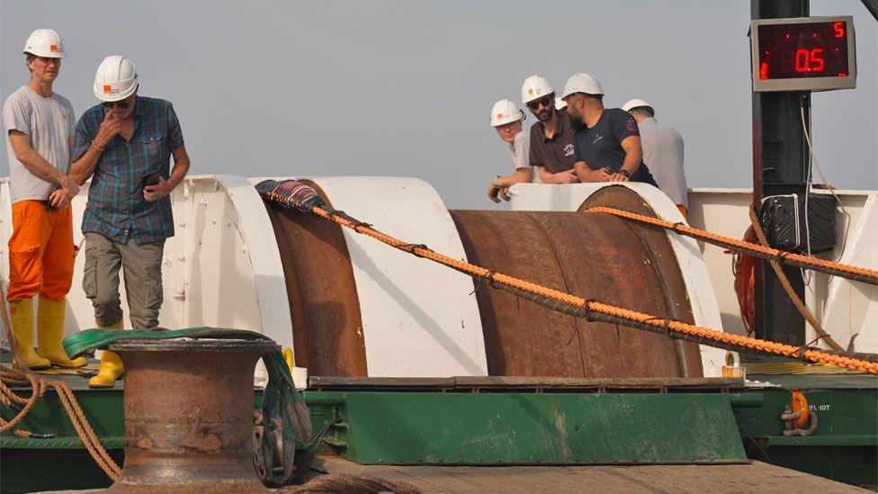 Several members of Léon Thévenin staff in white hardhats seen on deck near a giant pulley wheel used to lower internet cables.