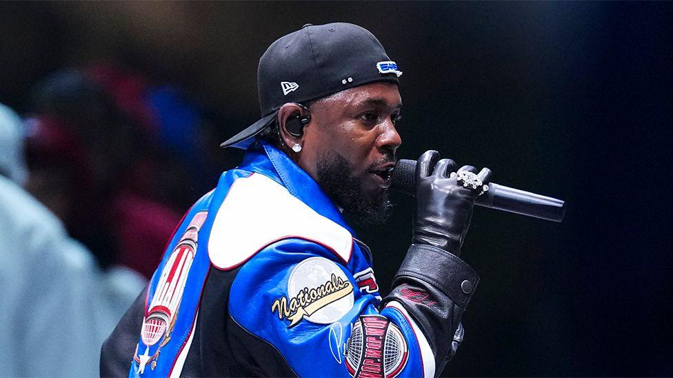 Kendrick Lamar, performing at the Super Bowl in New Orleans, wearing a black leather glove, and singing into a black microphone, with a reversed black cap.