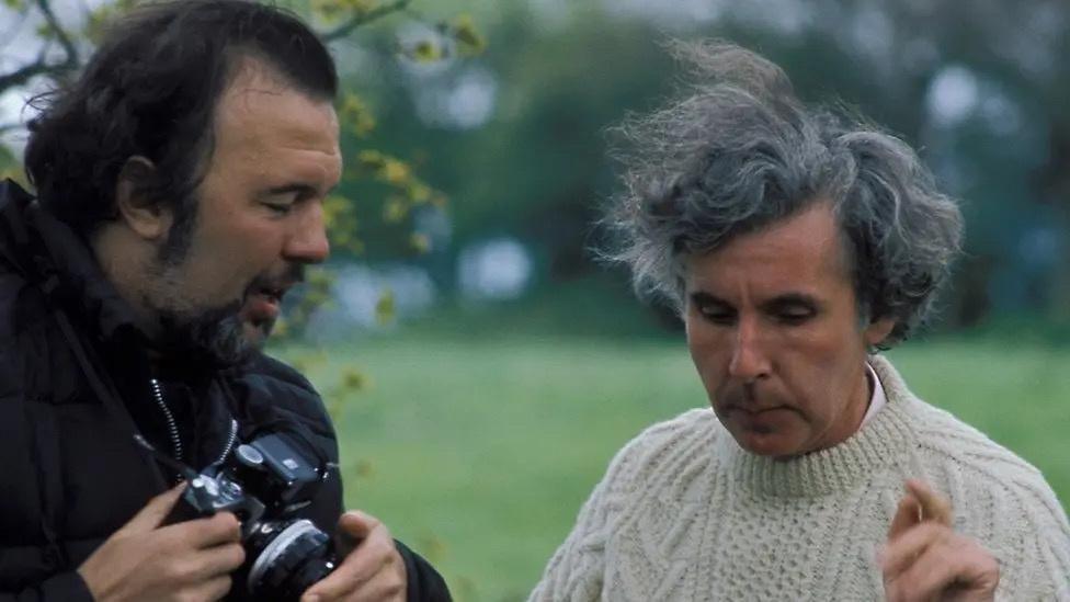 Sir Peter Hall, a dark-haired, bearded man wearing a dark coat and holding a stills camera, looks at Ronald Blythe who is wearing a white jumping and pointing with the forefinger of his left hand