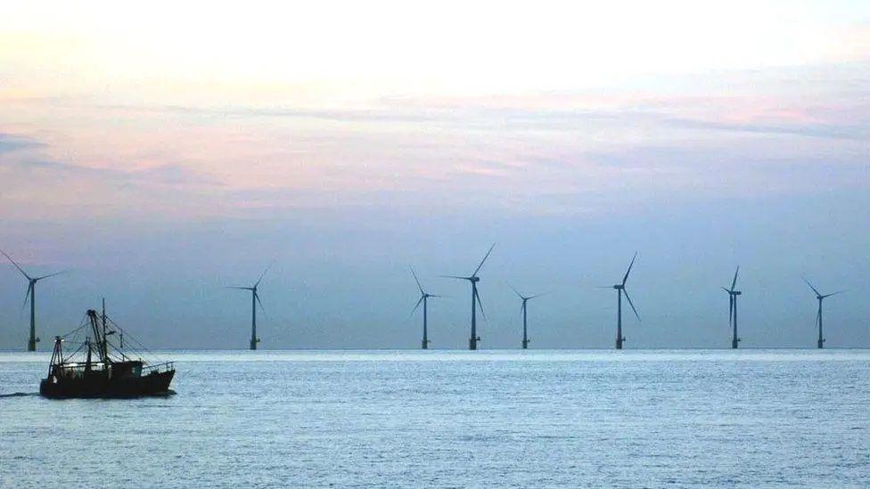 An offshore windfarm on the sea during sunset, with a fishing trawler in the foreground.