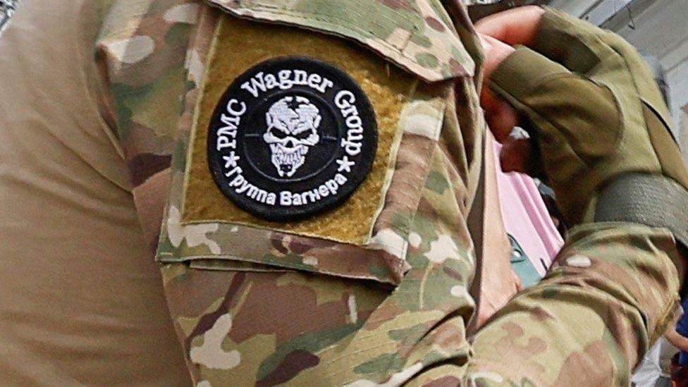 A close-up of a gloved hand and arm of a Wagner soldier, showing his camouflaged fatigues and the black and white Wagner logo featuring a white skull