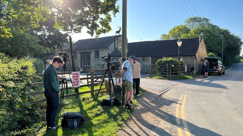 Multiple cameras on tripods are pointed over a fence at the side of the road. 