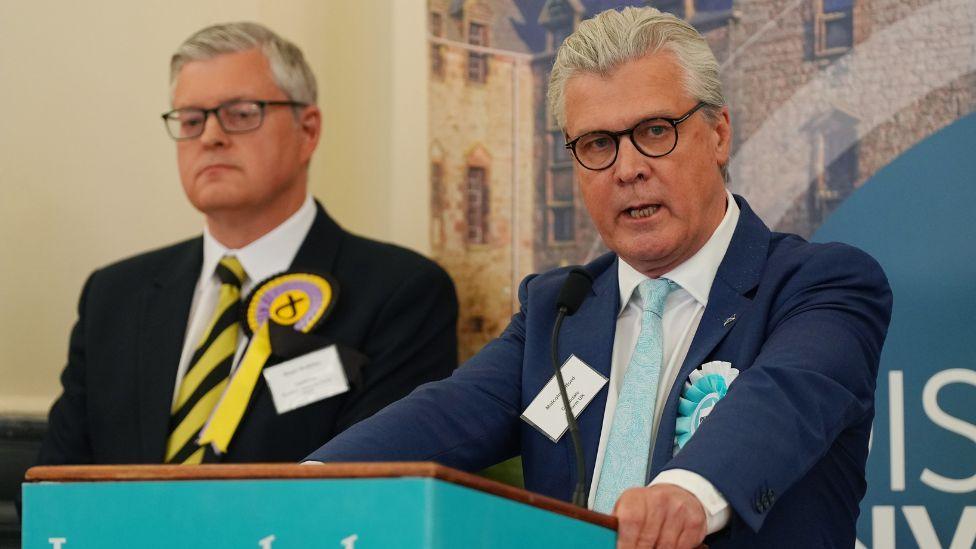 Malcolm Offord, i cili ka flokë gri dhe syze, flet në një podium me logon blu të zbehtë të Reform.