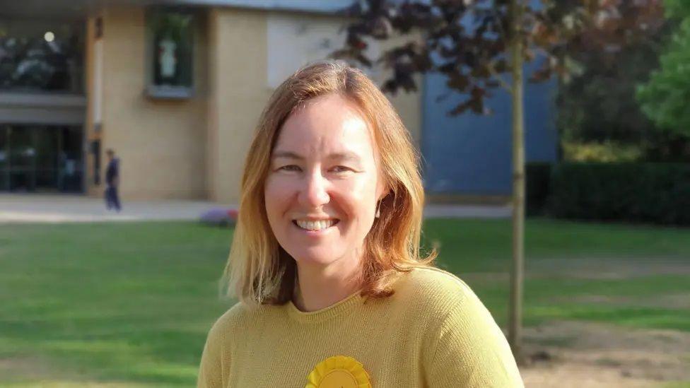 Marie Goldman standing outdoors in what looks like the garden of a block of flats, with a grass quad and tree behind her. She wears a yellow jumper and a yellow Lib Dem rosette. She has shoulder length blonde hair.