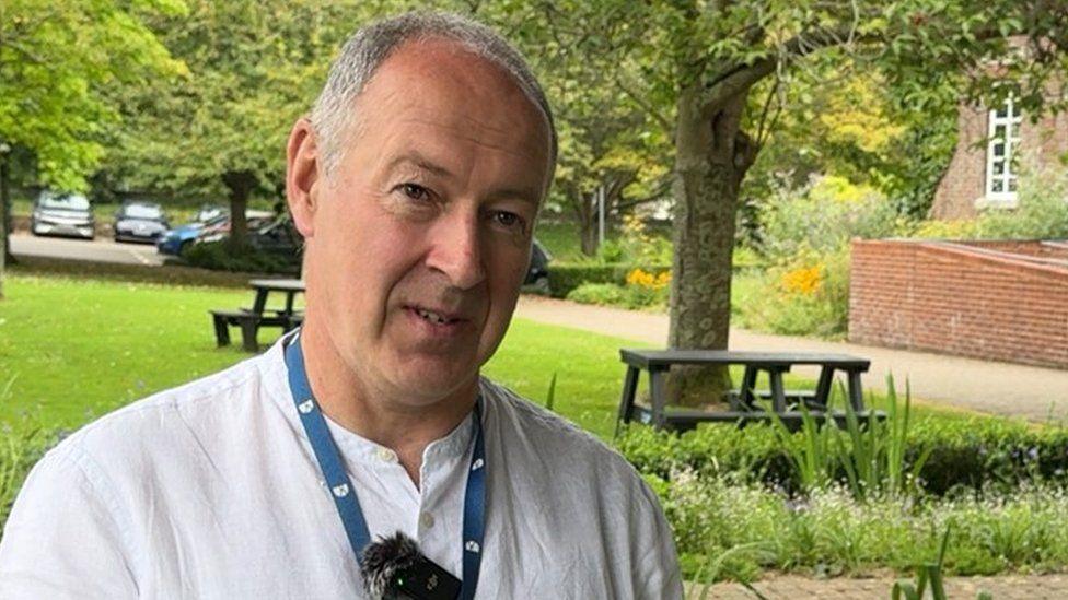 Nick Ireland in a white collarless shirt, standing in front of public gardens