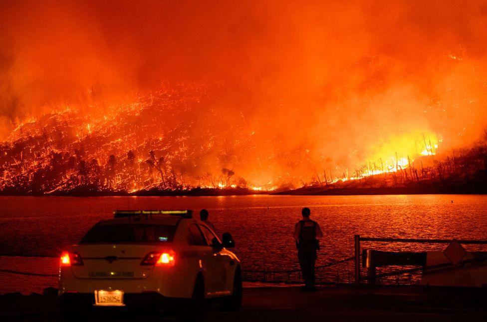 Law enforcement members watch as the Thompson fire burns over Lake Oroville in Oroville, California on July 2, 2024. A police car and officers are in the foreground, fire burns in the background.