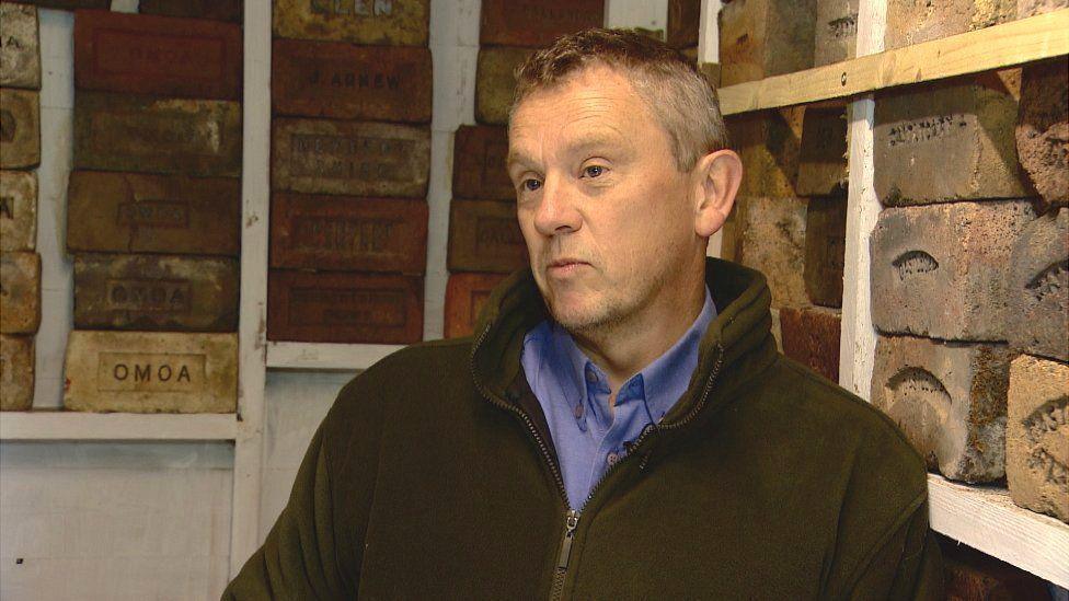 Mark Cranston is standing in front of shelves of bricks at his home in Jedburgh. He is wearing a brown fleece and blue shirt.