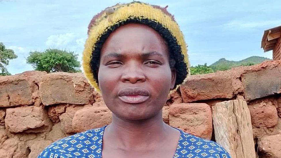 A close-up of HIV patient Eddah Simfukwe Banda who is wearing a yellow woolly hat and blue patterned top. She is standing against a roughly built brick wall with green trees and vegetation behind that. 