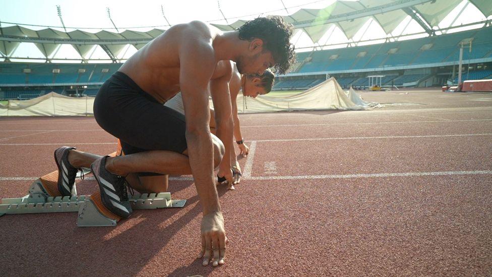 Abhay and Simran crouch at the starting line to practise the beginning of a race. He is in the foreground with black shorts and a bare torso, and she is behind him in white.