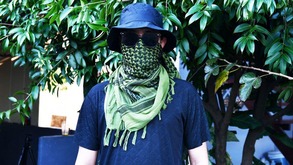 A man, disguised in sunglasses and with a green checked scarf over his face, wearing a blue bucket hat and a blue T-shirt