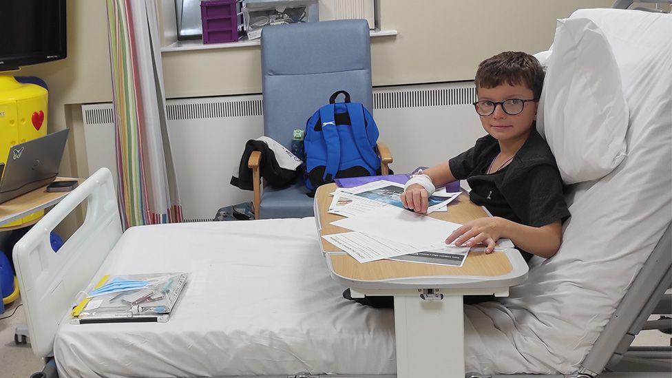 Dima Boichak sitting in a hospital bed after being given a transfusion of teplizumab. He is wearing glasses and a black top and writing on the table in front of him while sitting up in bed.