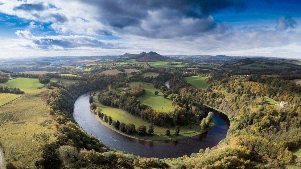 The river Tweed on a big bend in the foreground with hills in the distance. The banks are heavily wooded but there are also many fields visible 