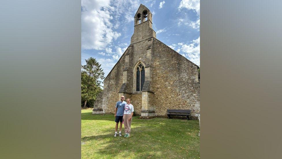 Kevin Todd and Sarah Carroll are standing on the grass in front of an old stone church. The church has a stained glass window and a bell tower at the top. Mr Todd is wearing a blue T-shirt and shorts. Ms Carroll is wearing a white T-shirt and brown trousers