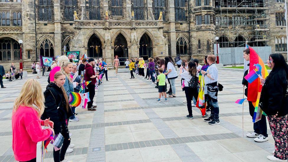 A drag queen, wearing a colourful dress and a bright red wig, walks between two lines of people, draped in rainbow flags and dressed in casual clothes, outside Rochdale's stone-built gothic town hall