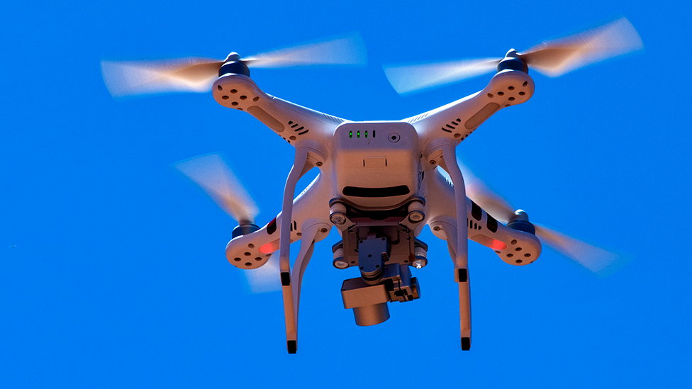 A generic image of a white domestic drone quadcopter in a blue sky. The drone is equipped with a silver camera underneath it