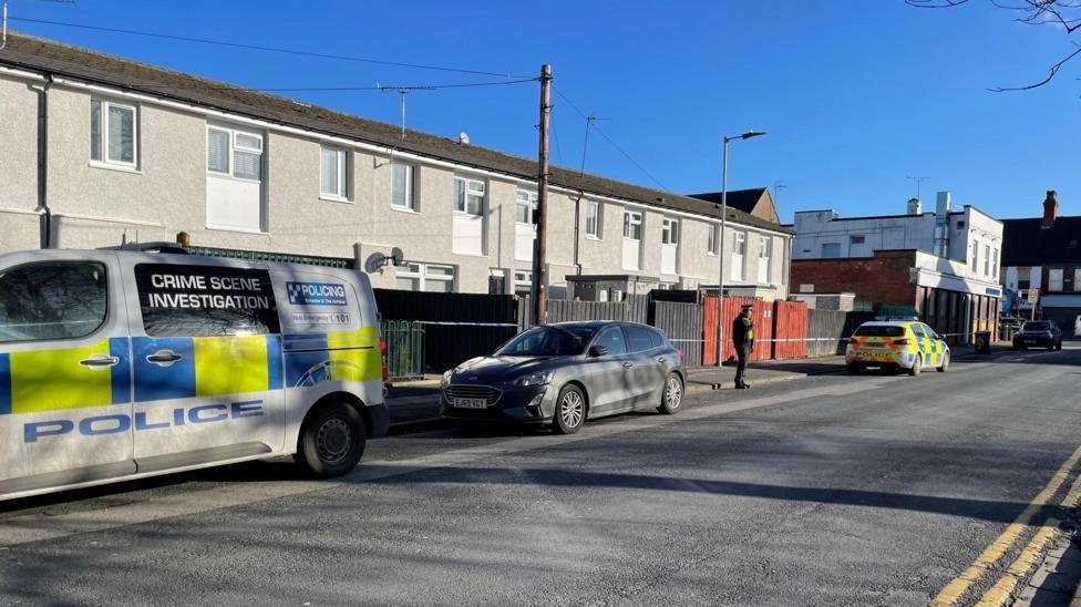 Barnsley Street in Hull. A row of grey terraced houses surrounded by police tape and vehicles. A large police van with the words "crime scene investigation" on the side is parked at the left of the image. A police officer, wearing black uniform, is also visible in the background.