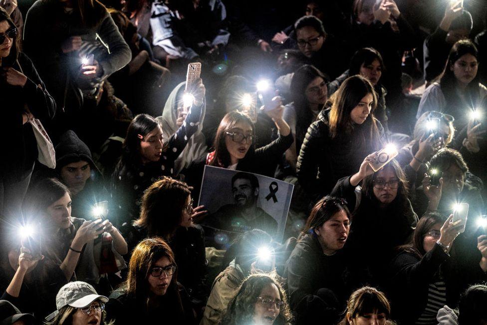 A group of young people light cell phones in the darkness of night. One person holds a large black and white picture of the singer Liam Payne