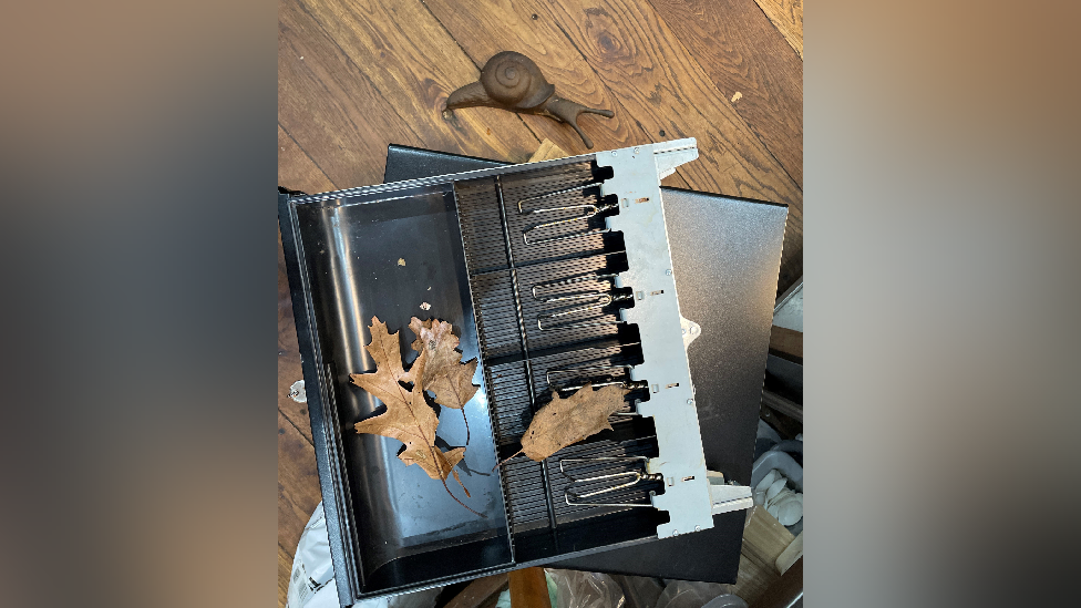 A broken till, the drawer is stacked on top of the main till with leaves and other fragments of debris inside the drawer. There is also a fake snail and a wooden floor in the background. 