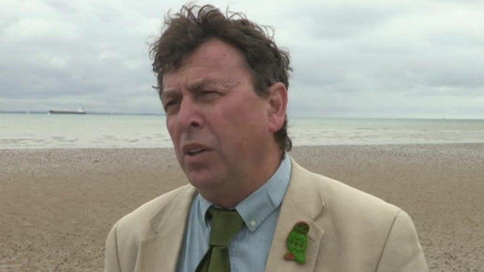 Michael Lilley gives an interview on a sand-coloured beach. He wears a beige jacket, blue shirt and green tie.