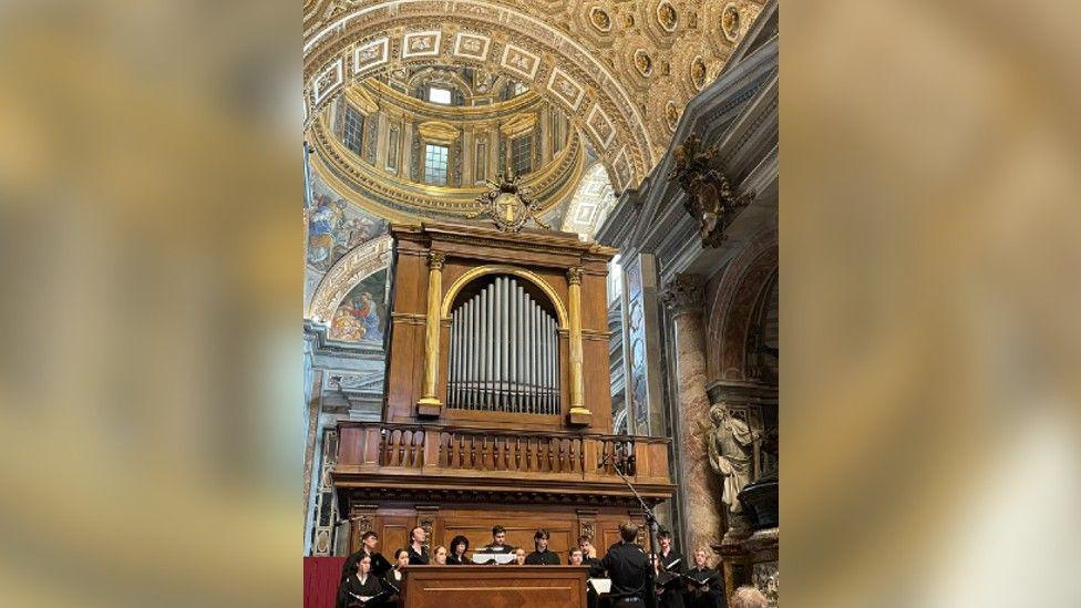 The choir singing at St Peter's Basilica. They are all dressed in black and there is an organ behind them. The dome is above them.