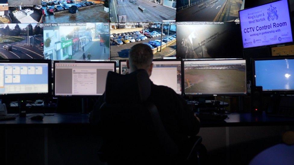 A man sits in front of a number of CCTV screens. His back is to the camera. It is dark. There are at least 14 screens in front of him.