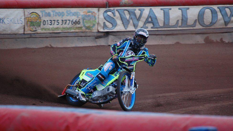 A speedway rider in action on a blue and neon yellow motorbike. The helmet and protective gear they are wearing matches their bike and also contains various logos from advertising companies. The rider looks to be skidding to a halt with dirt spraying to the left of the image from the back wheel of the bike. 