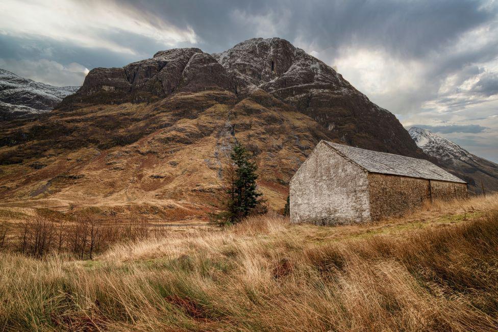 A stone shed is in the foreground surrounded by tall grass - a mountain rises up in the background.