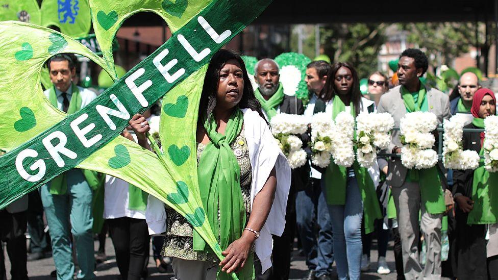 A person holds up a large green heart-shaped sign with the word 'Grenfell' on it, with other demonstrators in the background, as they walk to the Wall of Truth to mark the one year anniversary of the Grenfell fire in London on 14 June