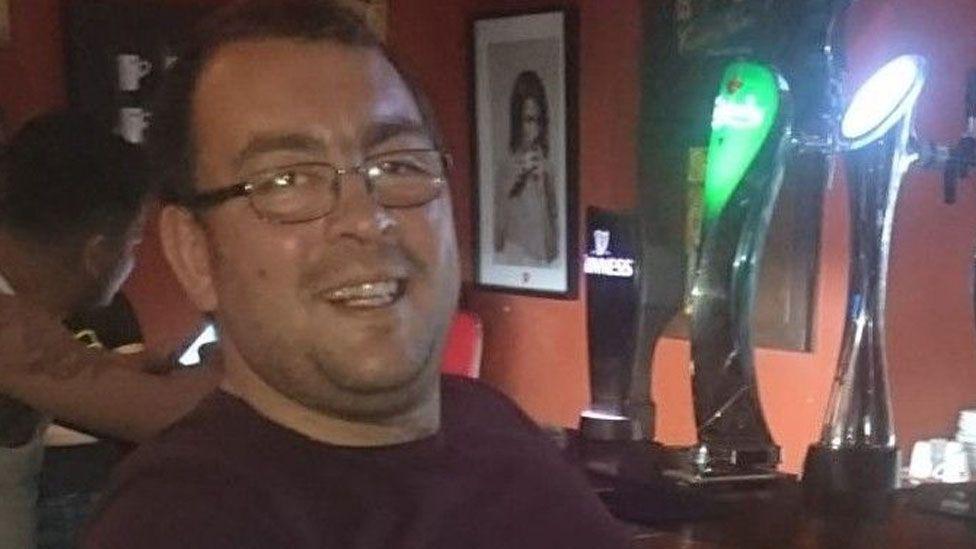 Head and shoulders of Danny Aitchison sat in a bar next to three pumps. He is smiling and has short dark hair and wire framed glasses and a burgundy tshirt