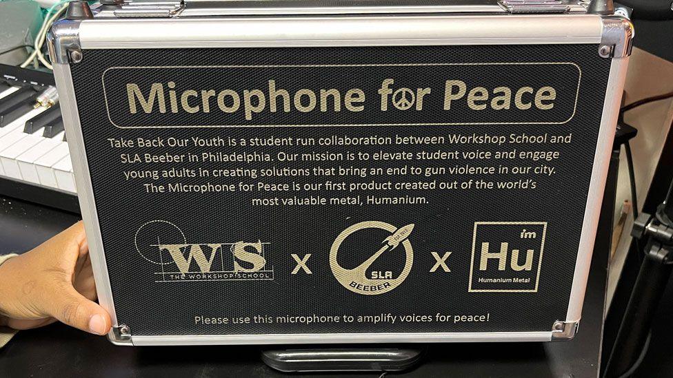 A close up of a black microphone box which says Microphone of Peace in white letters and underneath explains what the project is. It is being held up by a just seen hand on the left side.