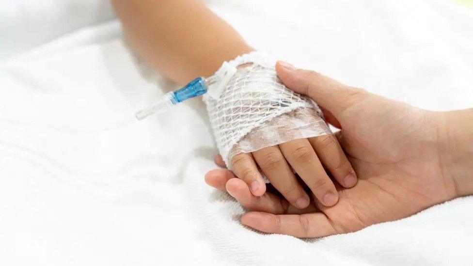 A child's hand, swathed in medical bandages, touches an adult's hand against what appears to be a white bedsheet.