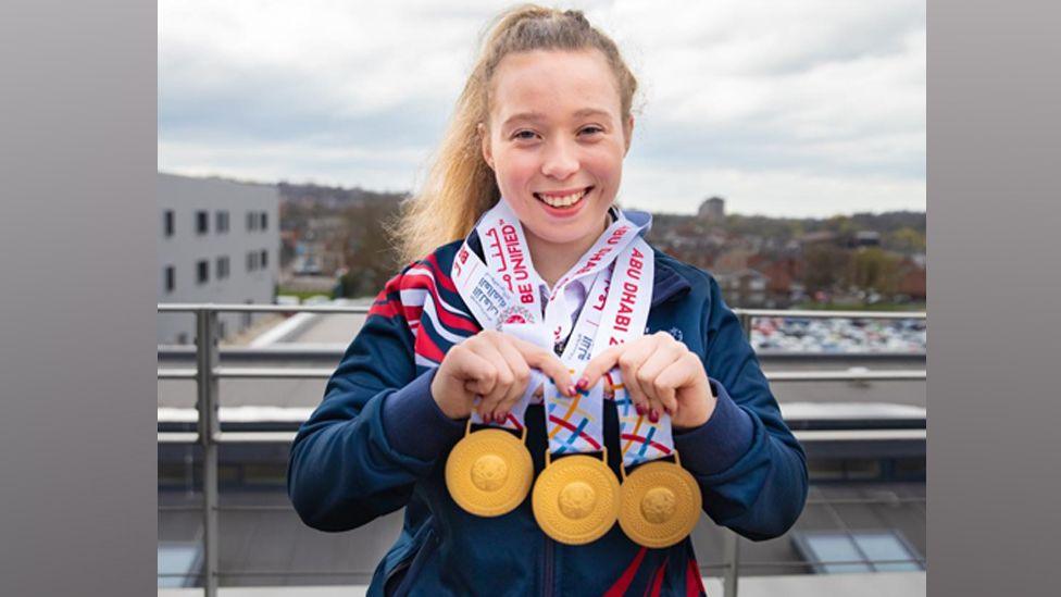Kiera Byland is smiling directly at the camera. Around her neck are three gold Special Olympics medals which she is holding up towards the camera.