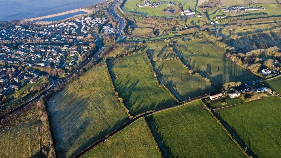 An aerial view of the land being developed fro new homes in Derry. The caw tounabout and foyle bridg, as well as existing homes can be seen, as  can a numbr of green fields.