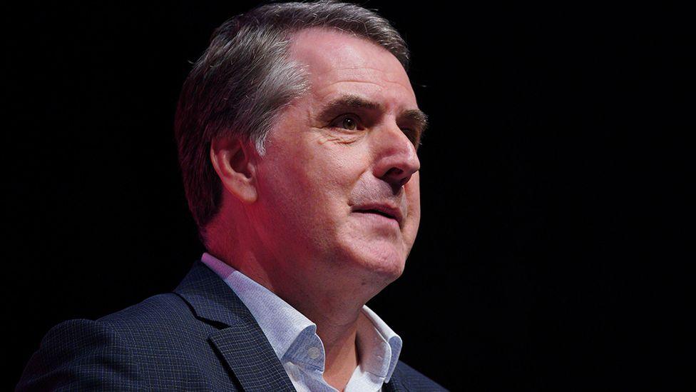 Head and shoulders image of Steve Rotheram, against a black background, looking to the right. He is wearing a blue suit and pale blue open-neck shirt.