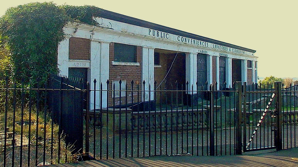 Flat-roof white toilet block photographed from the street with its doors and windows boarded up