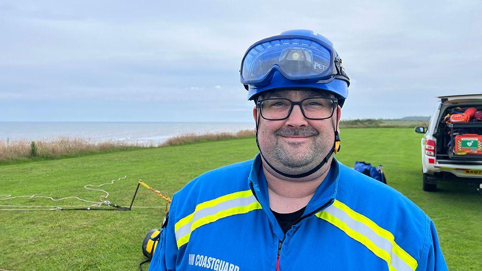 Ben Colombari looking directly into the camera stood on the clifftop in Hornsea wearing a blue coastguard uniform with a blue helmet and glasses