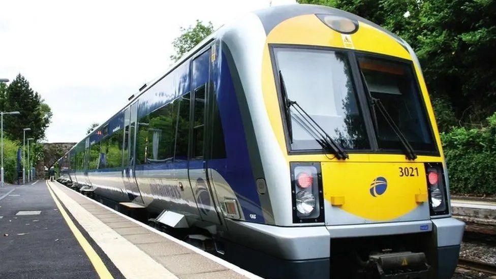 A Translink train stopped at a station in Belfast. The train is blue, yellow and grey and is pictured from the front at a slight angle. 