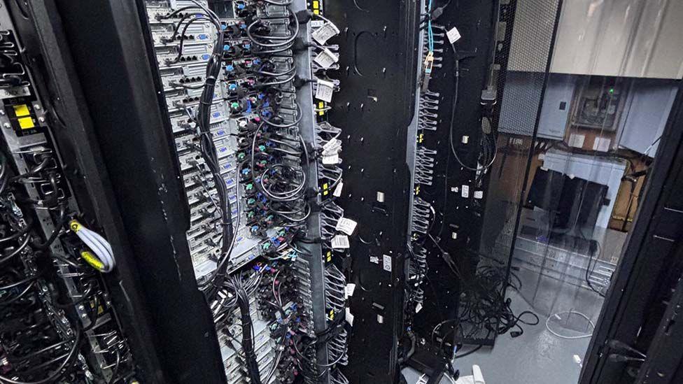 A row of computer servers, tall stacked black computer cabinets with multiple trays and wire connections