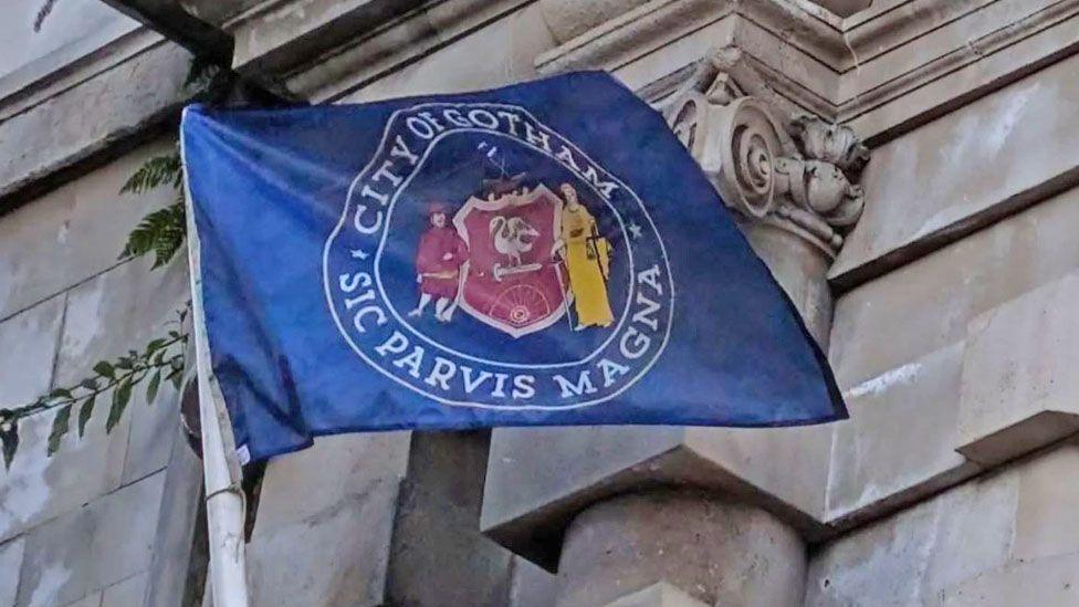 A blue flag is flying from a pole on the side of a building in Liverpool. In white writing around a red and yellow crest it says City of Gotham, Sic Parvis Magna.