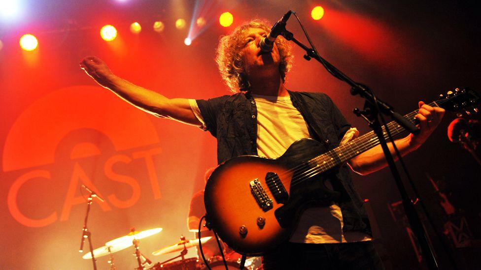 Cast's John Power sings into a microphone and plays a guitar in front of a drummer and a large backdrop with the word "Cast" on it