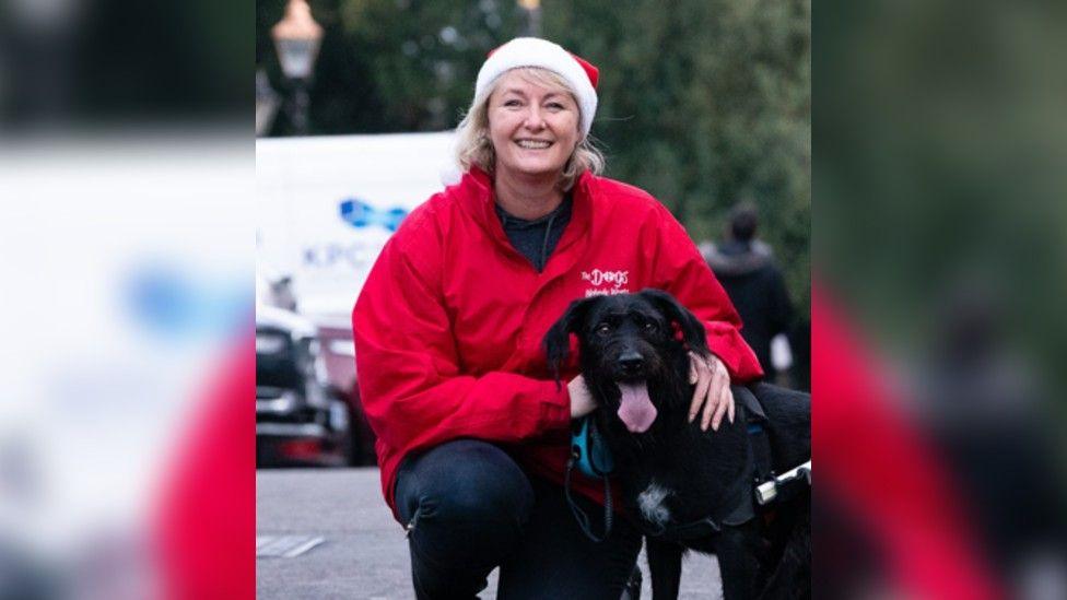 Jo Drury is kneeling down stroking a black labrador, she is wearing a red sweatshirt, dark jeans and a santa's hat.