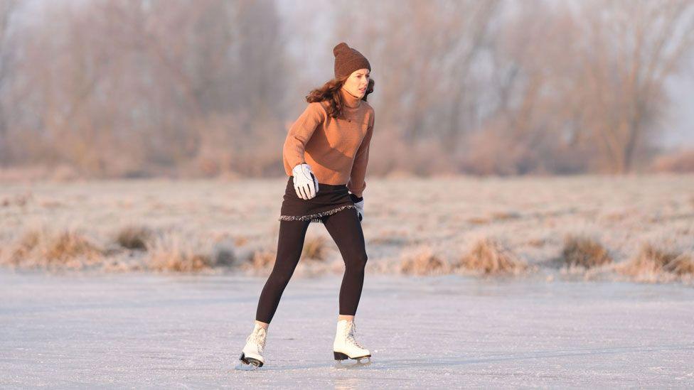 A woman skating at speed on a frozen field at Upware, Cambridgeshire. She is wearing a brown bobble hat and leggings, an orange jumper and white padded gloves and ice skating boots. Behind her is the field edge and eyond it wintery trees